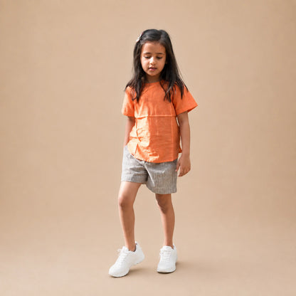 A young girl stands against a solid beige studio background wearing a bright orange short-sleeve top, light gray patterned shorts, and white sneakers.