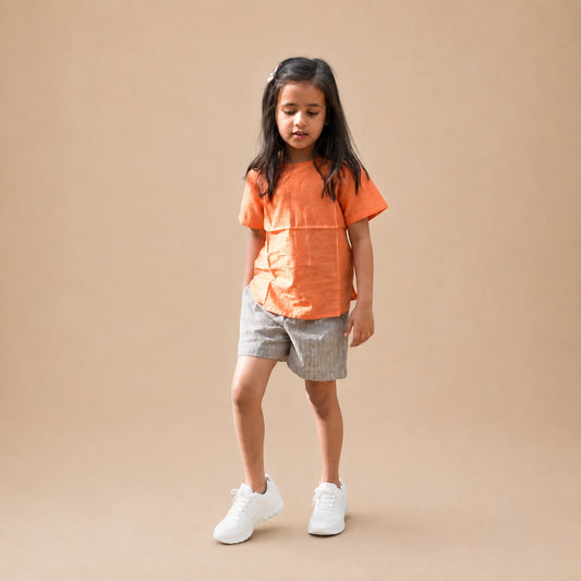A young girl stands against a solid beige studio background wearing a bright orange short-sleeve top, light gray patterned shorts, and white sneakers.