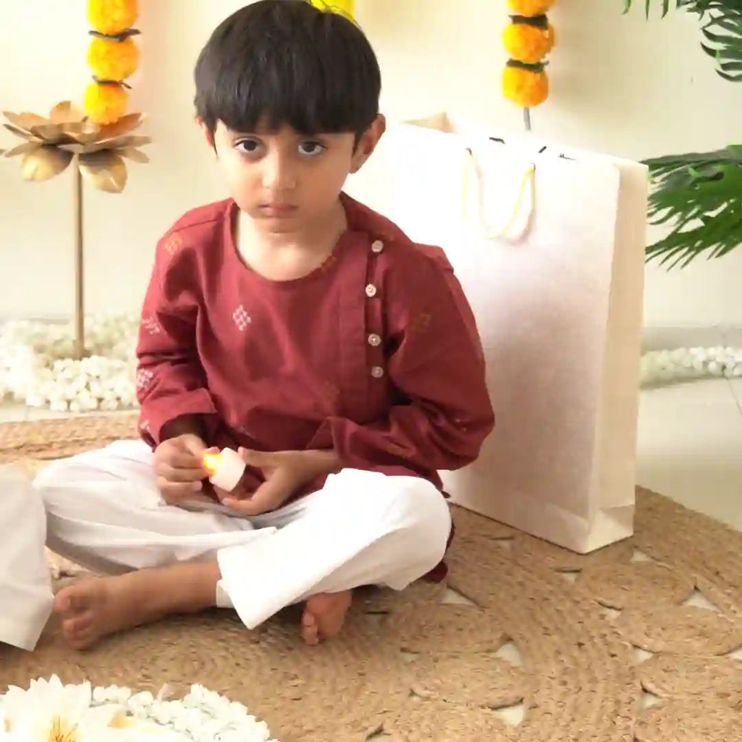 Boy in a maroon kurta with an angled button placket and white pajama sitting on a jute mat holding a tea light