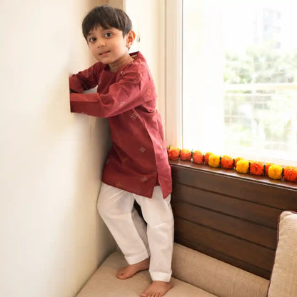Boy in a maroon handloom cotton kurta and white pajama standing by a window with a marigold garland