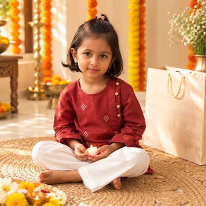 Girl in a maroon handloom cotton kurta and white pajama sitting cross-legged on a jute mat holding a lit diya