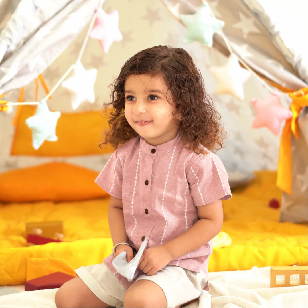 Smiling child in a mauve handloom cotton shirt with white raindrop stripes and wooden buttons, sitting in a play tent