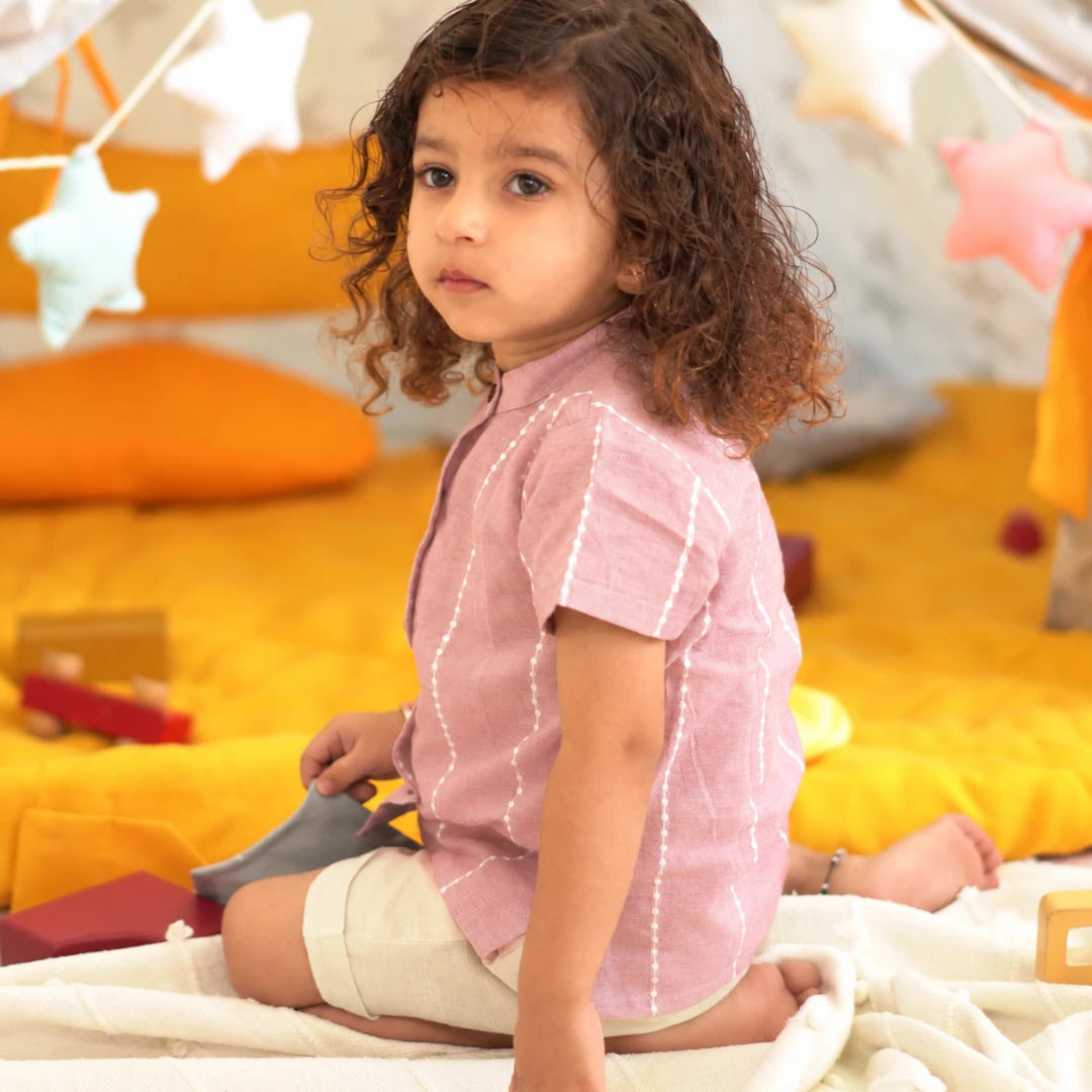 Side view of a child in a relaxed-fit mauve cotton shirt with mandarin collar and beige shorts, sitting on a soft blanket