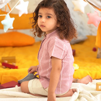 Side view of a child in a relaxed-fit mauve cotton shirt with mandarin collar and beige shorts, sitting on a soft blanket