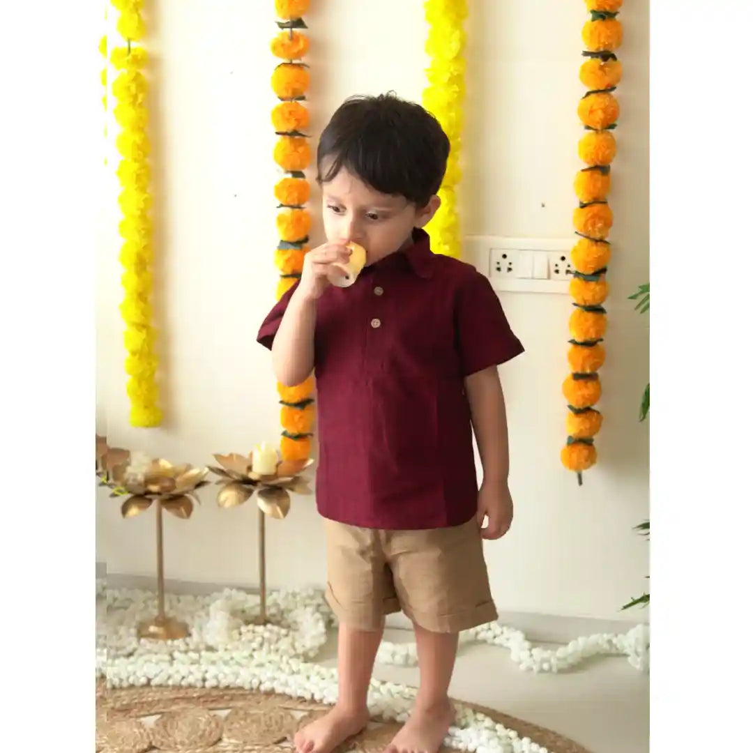 Boy in a maroon polo neck shirt and beige shorts standing by marigold garlands and gold lotus candle holders