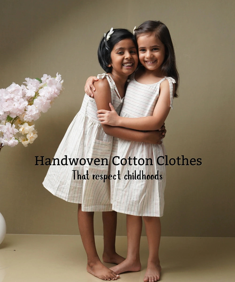 Two girls hugging in white handwoven cotton dresses, with the text handwoven cotton clothes that respect childhoods