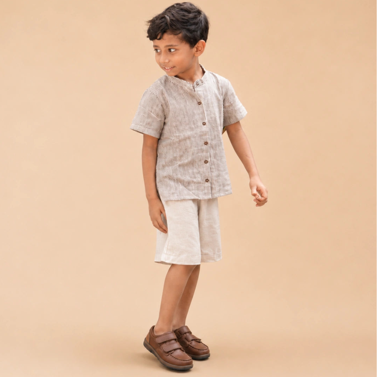 A young boy modeling a stripe short-sleeve shirt and shorts in handwoven cotton paired with brown leather shoes, against a solid beige studio background.
