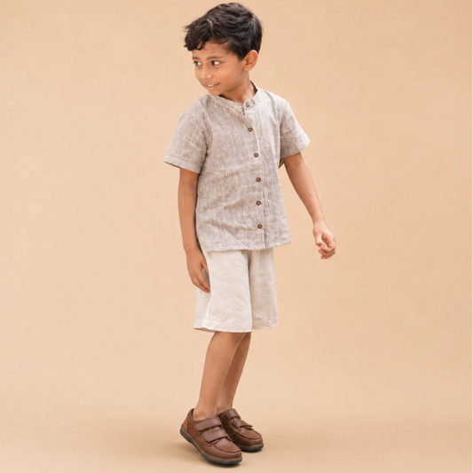 A young boy modeling a stripe short-sleeve shirt and shorts in handwoven cotton paired with brown leather shoes, against a solid beige studio background.