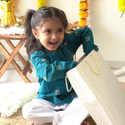 Girl in a teal handloom cotton kurta and white pajama smiling as she opens a festive gift bag
