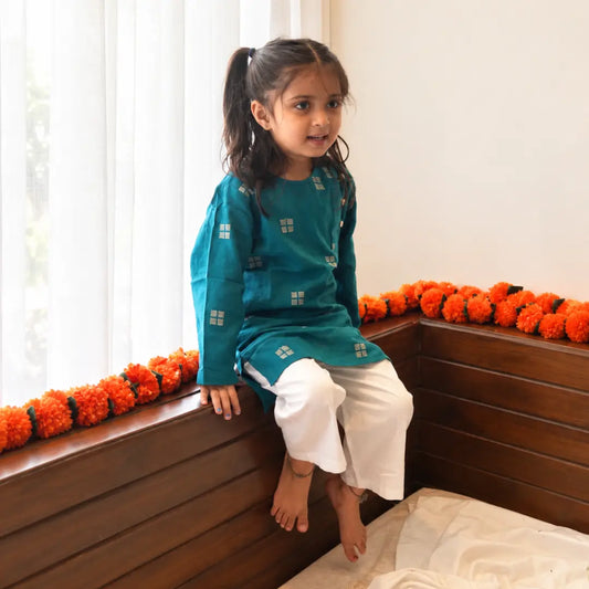 Girl in a teal handloom cotton kurta and white pajama sitting by a window with marigold garlands