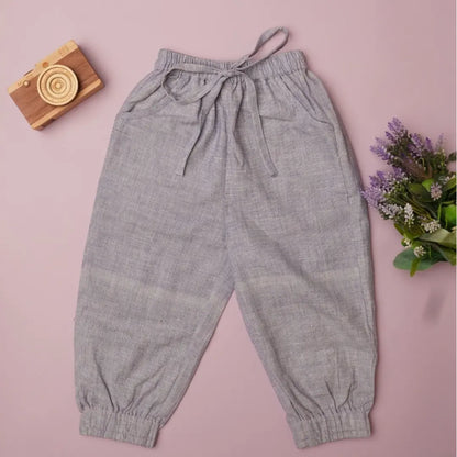 Light purple textured cottonjogger pants with an elastic drawstring waist and elastic cuffs, laid flat on a dusty pink background with a wooden camera and purple flowers as props.